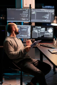 Web developer comparing data on papers and multple screens, working late at night on IT programming analysis. Coder looking at html script and information on clipboard files in office.