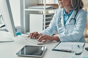 Close up of female doctor in white lab coat working using computer while sitting in her office